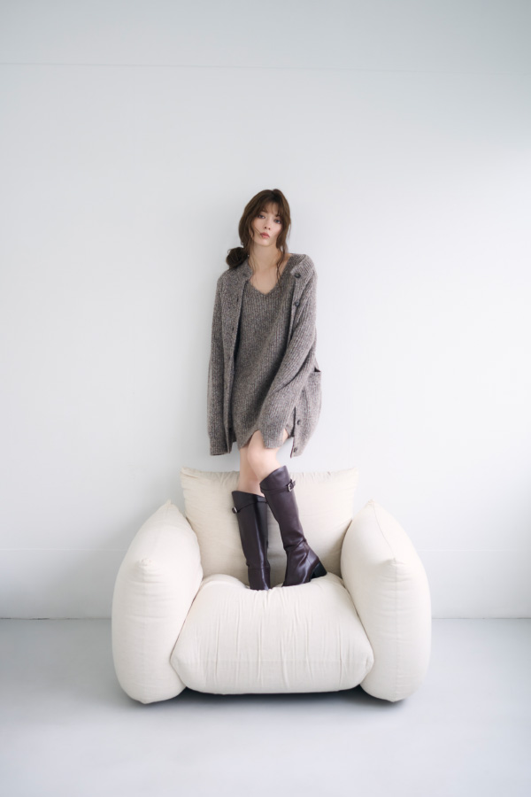 ロングカーデ＆ミニニットワンピｓｅｔとバックルストラップロングブーツを着用して白いソファーに立てる女性