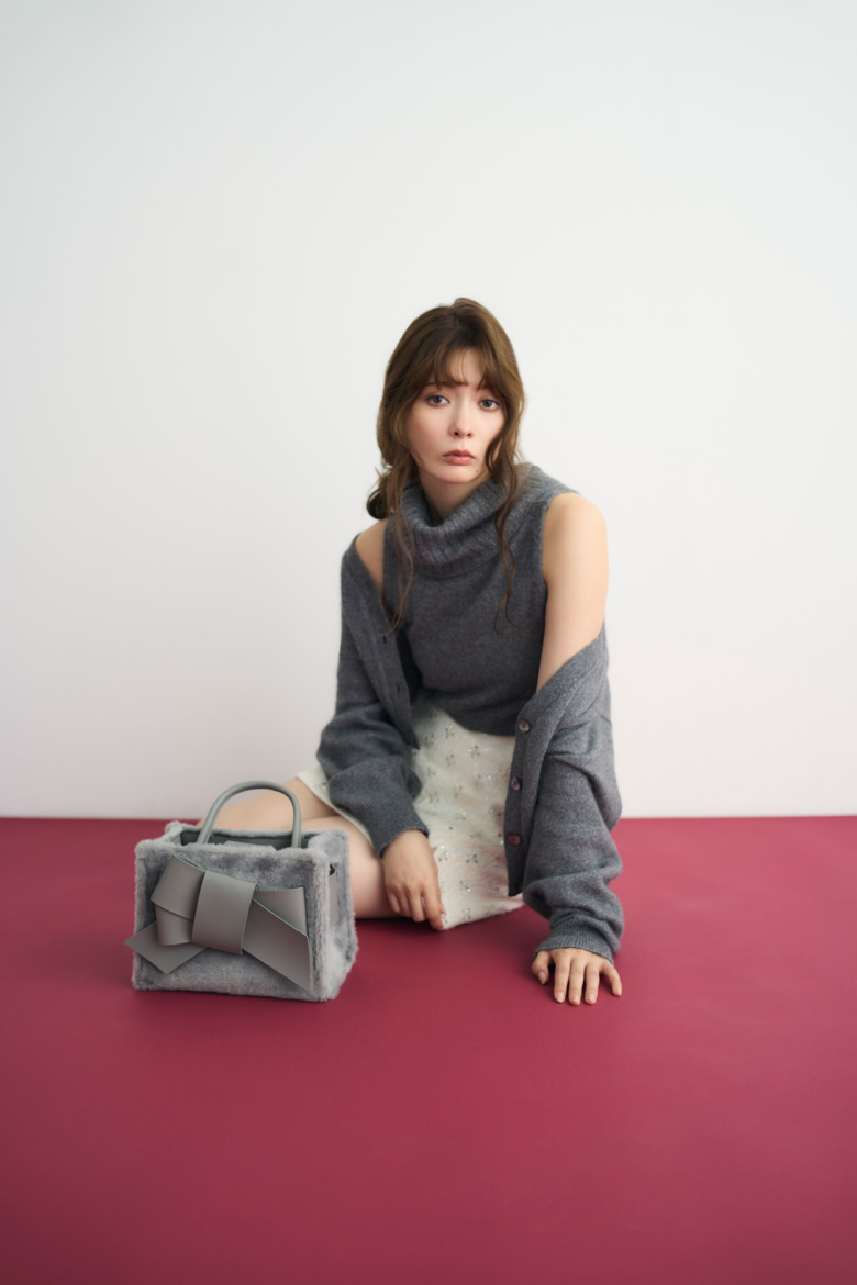 グレイ色のラクーンタートルカーデアンサンブルニットと【Holiday】ファーリボンミドルバッグ、マトラッセラメＪＱミニスカートを着てバーガンディー色の床に座ってる女性