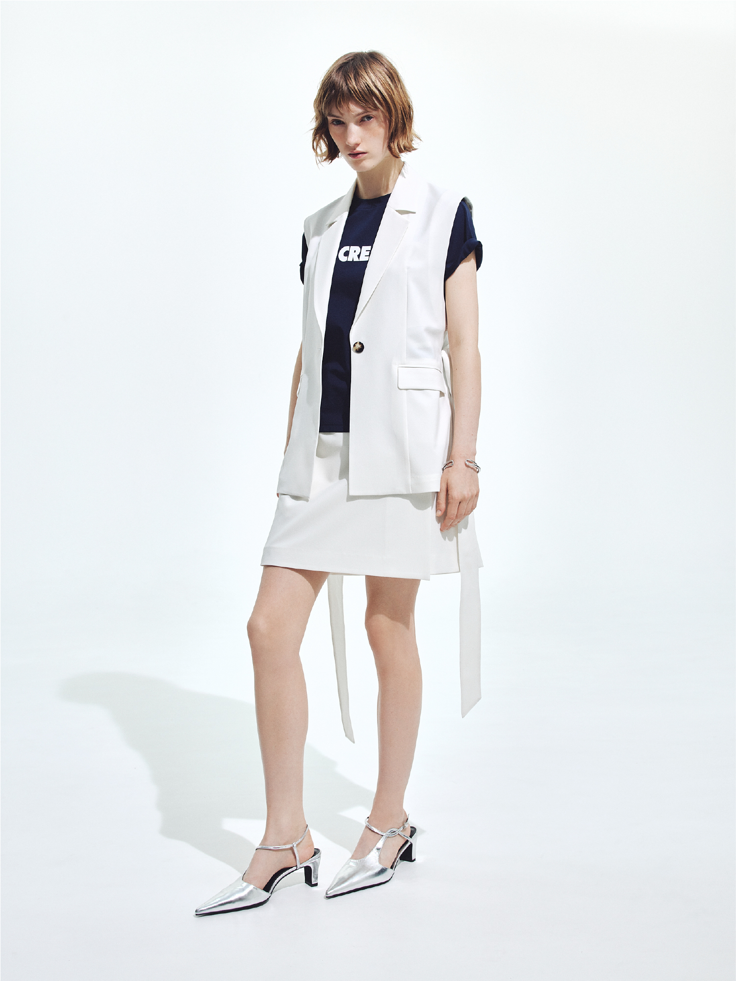 ボックスプリーツジレ・ロゴTシャツ・ウエストバックルミニスカート・トーションナローストラップミュール・ニュアンスクロスバングルを着用した女性