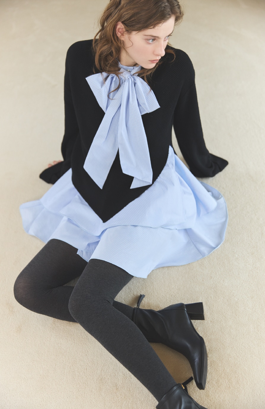 ブラックのリボンタイニットとライトブルーのチュニックが合体したコンビドレスを着た女性がアイボリーのリボンミドルバッグを持っている写真