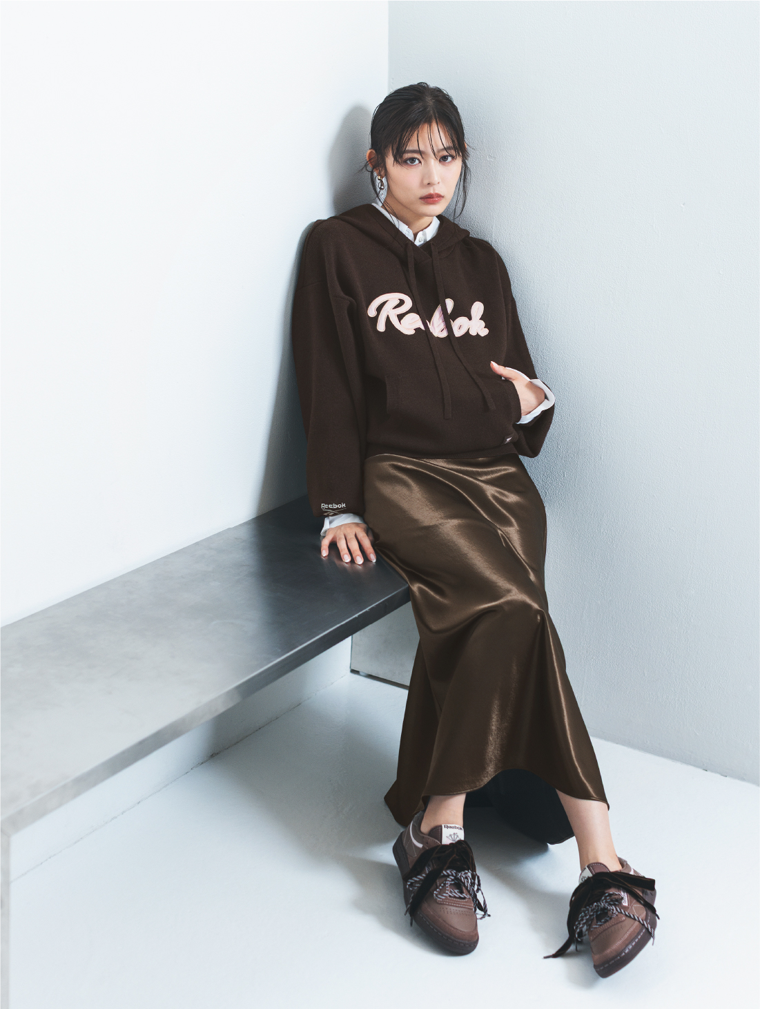 【Reebok】サテンロゴニットフーディー・ハイウエストサテンナロースカート・カメリア スタンダードシャツ・【Reebok】スニーカー・2WAYトリニティドロップピアスを着用した女性